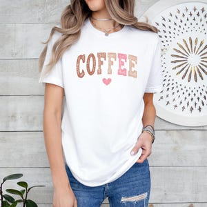 May include: White t-shirt with the word "COFFEE" printed in brown, pink, and white with a pink heart underneath.