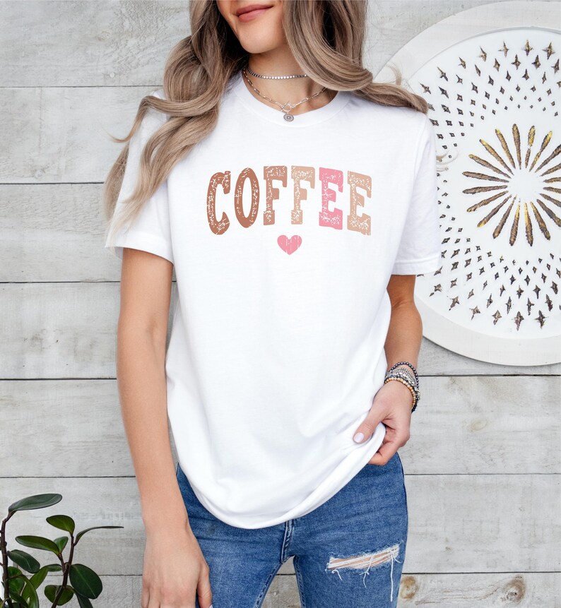 May include: White t-shirt with the word "COFFEE" printed in brown, pink, and white with a pink heart underneath.
