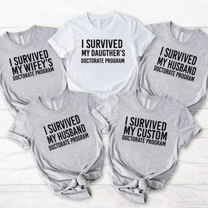 May include: Five grey and white t-shirts with black text that reads "I survived my [relationship] doctorate program". The relationships are wifey, daughter, husband, husband, and custom.