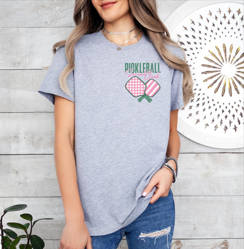 Op de afbeelding: Grijs T-shirt met een afbeelding van twee roze en witte pickleball-rackets met de tekst "Pickleball Granny Club".