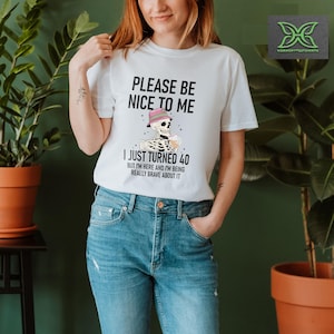 May include: White t-shirt with the text "PLEASE BE NICE TO ME I JUST TURNED 40 BUT I'M HERE AND I'M BEING REALLY BRAVE ABOUT IT." The graphic features a skeleton wearing a hat and holding a drink. The shirt is worn by a person.