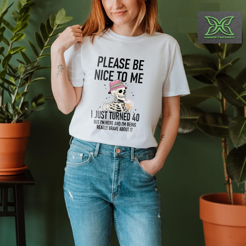 May include: White t-shirt with the text "PLEASE BE NICE TO ME I JUST TURNED 40 BUT I'M HERE AND I'M BEING REALLY BRAVE ABOUT IT." The graphic features a skeleton wearing a hat and holding a drink. The shirt is worn by a person.