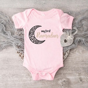 May include: Pink baby onesie with a black crescent moon and the text "my first Ramadan" in gold glitter.