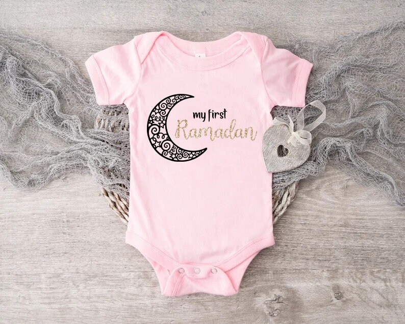 May include: Pink baby onesie with a black crescent moon and the text "my first Ramadan" in gold glitter.