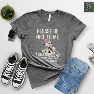 May include: A gray t-shirt with the text "PLEASE BE NICE TO ME" and "I JUST TURNED 40". The graphic shows a skeleton wearing a hat and holding a drink. Black sneakers and blue jeans are also visible.