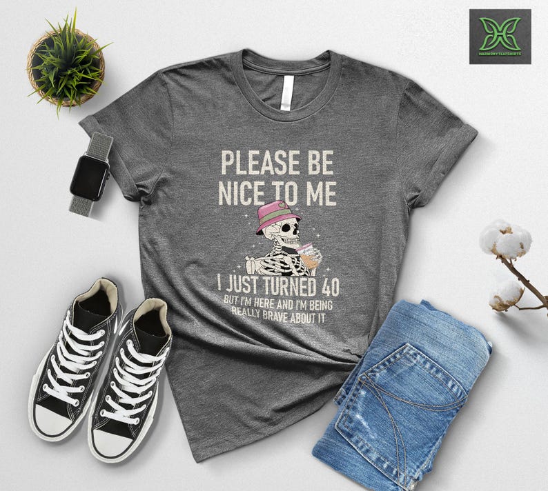 May include: A gray t-shirt with the text "PLEASE BE NICE TO ME" and "I JUST TURNED 40". The graphic shows a skeleton wearing a hat and holding a drink. Black sneakers and blue jeans are also visible.