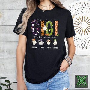 Op de afbeelding: Zwart T-shirt met de tekst "This Gigi loves her spooky crew" in een kleurrijk Halloween-thema. Het ontwerp bevat geesten en de namen Olivia, Emily, Noah en Sophie.