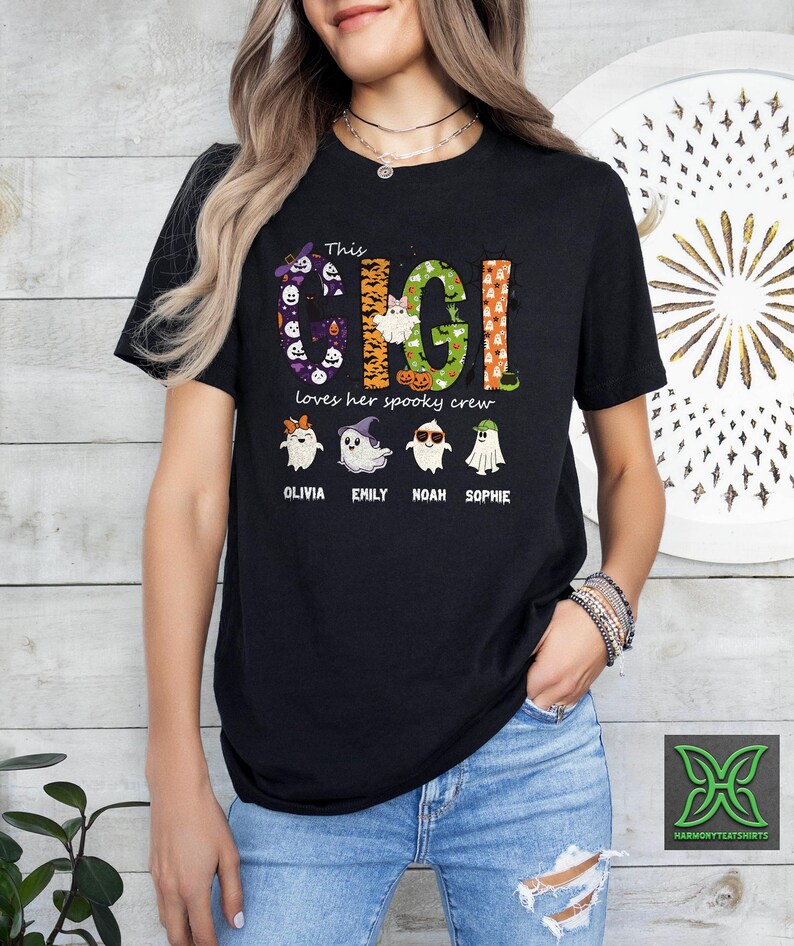 Op de afbeelding: Zwart T-shirt met de tekst "This Gigi loves her spooky crew" in een kleurrijk Halloween-thema. Het ontwerp bevat geesten en de namen Olivia, Emily, Noah en Sophie.