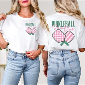 Op de afbeelding: Twee witte T-shirts met een roze en groen pickleball-paddle ontwerp en de tekst "Pickleball Social Club".