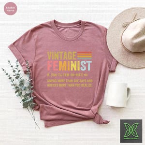 May include: A heather mauve t-shirt with the text "Vintage Feminist" in rainbow colors. The text "N. [VIN-TIJ FEM-UH-NIST]" is below the word "Feminist" and the text "KNOWS MORE THAN SHE SAYS AND NOTICES MORE THAN YOU REALIZE." is below that.