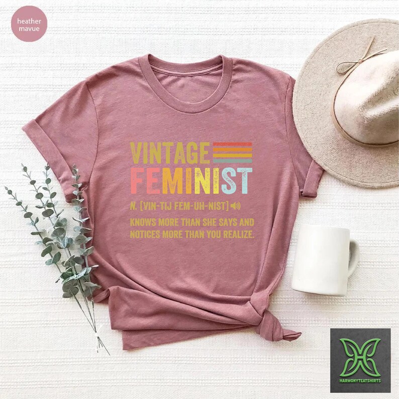 May include: A heather mauve t-shirt with the text "Vintage Feminist" in rainbow colors. The text "N. [VIN-TIJ FEM-UH-NIST]" is below the word "Feminist" and the text "KNOWS MORE THAN SHE SAYS AND NOTICES MORE THAN YOU REALIZE." is below that.