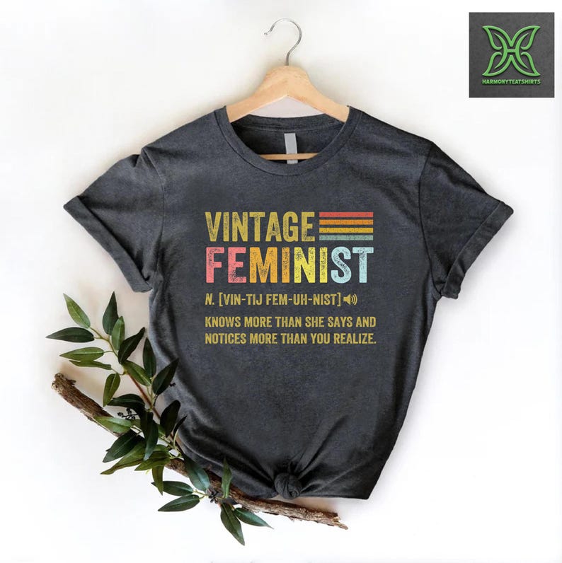 May include: A dark gray t-shirt with a vintage-style graphic that reads "Vintage Feminist" with a rainbow stripe design. The text below the graphic defines the word "Feminist" as "[VIN-TIJ FEM-UH-NIST]" and states that a feminist "knows more than she says and notices more than you realize."