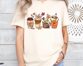 May include: A white t-shirt with a graphic of four fall-themed coffee cups. The cups are decorated with autumn leaves, a bow, and the text "Pumpkin Spice".