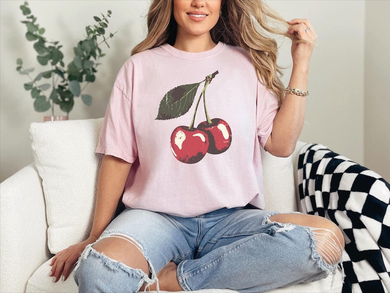 May include: A light pink t-shirt featuring a graphic of two red cherries with a green leaf. The shirt is worn with blue jeans. The cherries have a glossy appearance. The shirt is a casual style.