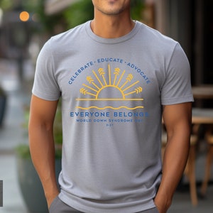 May include: A gray t-shirt with a blue and yellow graphic design. The graphic features a stylized sun with rays and the text "Celebrate, Educate, Advocate. Everyone Belongs. World Down Syndrome Day 3-21".