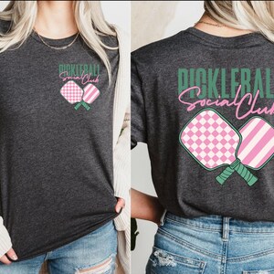 Op de afbeelding: Een donkergrijs T-shirt met een roze en groen pickleball-paddle ontwerp en de tekst "Pickleball Social Club" in groen.