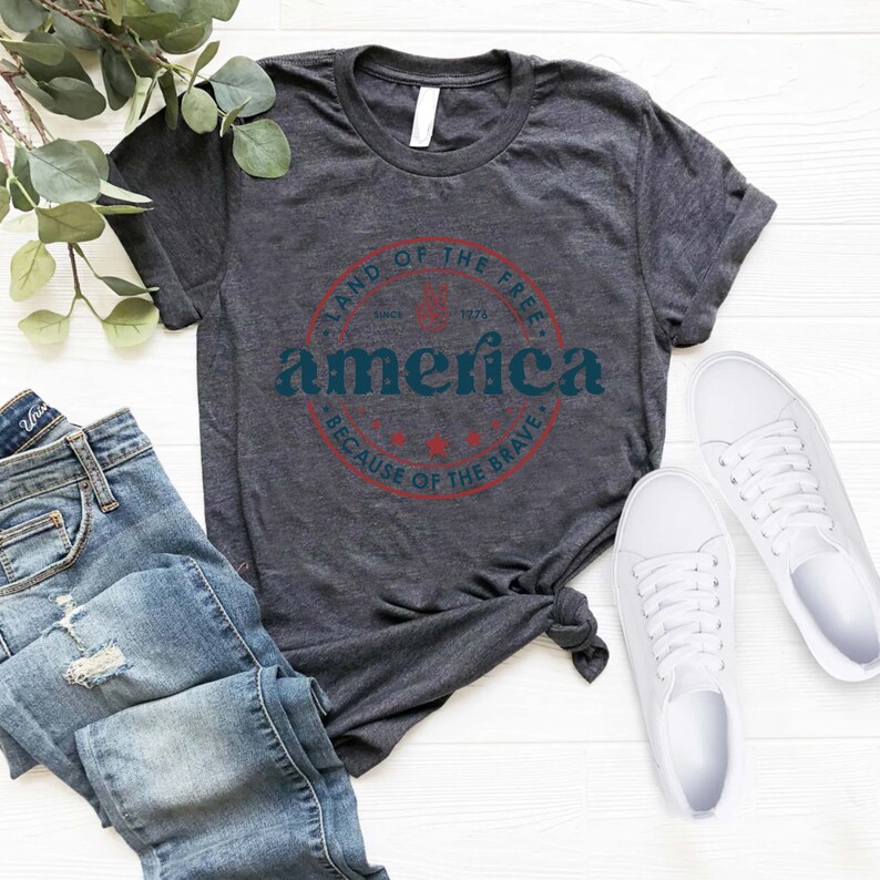 May include: A dark grey t-shirt with a red, white, and blue circular graphic that says "Land of the Free, Since 1776, America, Because of the Brave".