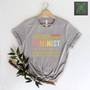 May include: Gray t-shirt with a vintage-style graphic that reads "Vintage Feminist" and a definition of the word. The graphic is in rainbow colors.