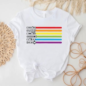 Puede incluir: Camiseta blanca con un dise&ntilde;o de arco&iacute;ris con sables de luz. Los colores del arco&iacute;ris son rojo, naranja, amarillo, verde, azul y morado.