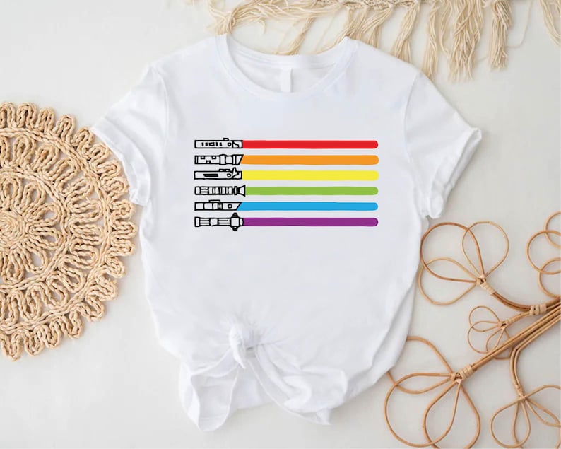 Puede incluir: Camiseta blanca con un dise&ntilde;o de arco&iacute;ris con sables de luz. Los colores del arco&iacute;ris son rojo, naranja, amarillo, verde, azul y morado.