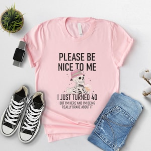May include: A light pink t-shirt with the text "PLEASE BE NICE TO ME" and "I JUST TURNED 40". The graphic shows a skeleton wearing a hat and holding a drink. Black high-top sneakers and blue jeans are also in the image.