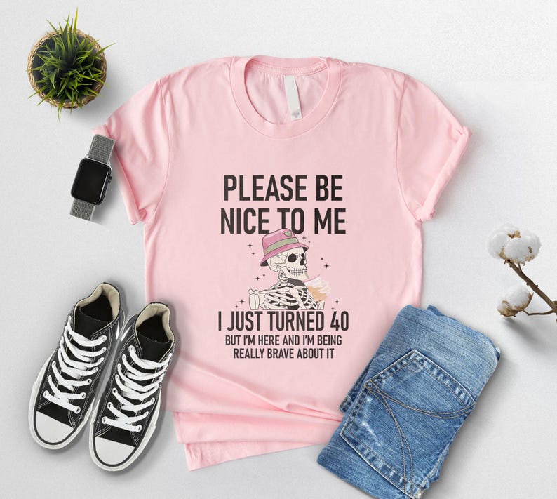 May include: A light pink t-shirt with the text "PLEASE BE NICE TO ME" and "I JUST TURNED 40". The graphic shows a skeleton wearing a hat and holding a drink. Black high-top sneakers and blue jeans are also in the image.