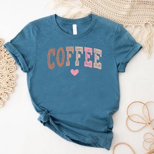 May include: A teal blue t-shirt with the word "COFFEE" printed in pink, brown, and white letters. A small pink heart is printed below the word.
