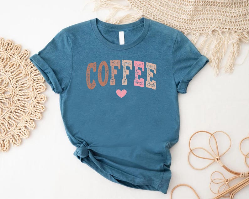 May include: A teal blue t-shirt with the word "COFFEE" printed in pink, brown, and white letters. A small pink heart is printed below the word.