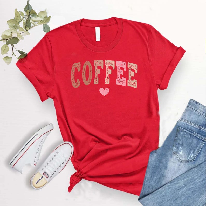 May include: Red t-shirt with the word "COFFEE" in pink, brown, and white letters with a small pink heart underneath. The shirt is rolled up at the sleeves and is laying on a white surface with a pair of white sneakers and blue jeans.