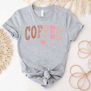 May include: A gray t-shirt with the word "COFFEE" printed in brown, pink, and peach colors. A small pink heart is below the word.