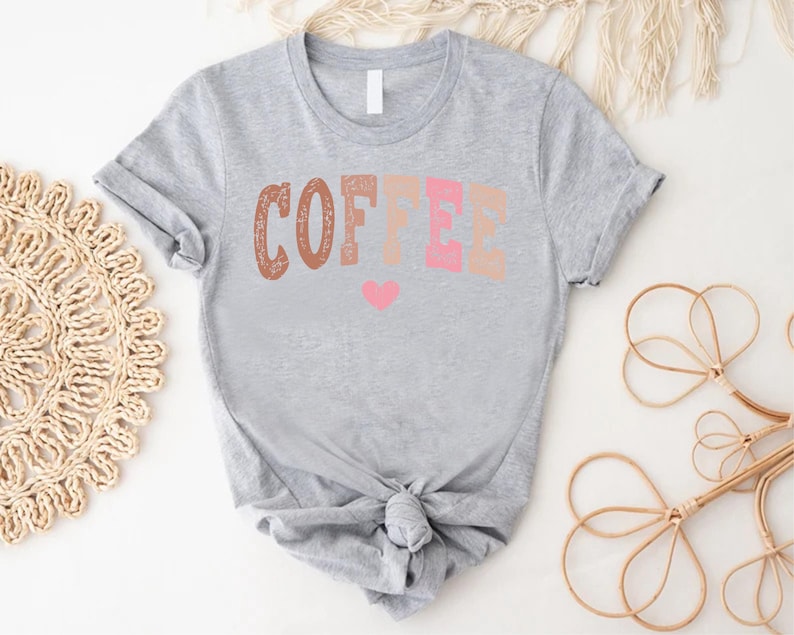 May include: A gray t-shirt with the word "COFFEE" printed in brown, pink, and peach colors. A small pink heart is below the word.