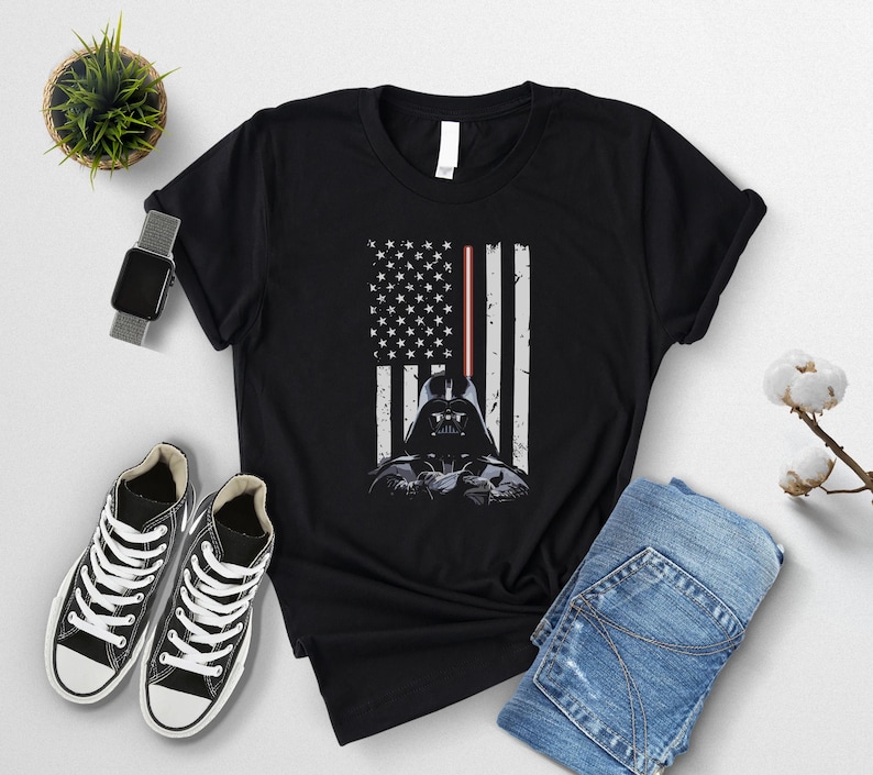 Puede incluir: Camiseta negra con un dise&ntilde;o gr&aacute;fico del casco de Darth Vader integrado en una bandera estadounidense. Las rayas de la bandera son blancas y el sable de luz es rojo. La camiseta se muestra con zapatillas negras y vaqueros azules.