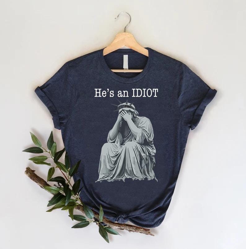 May include: Dark blue t-shirt featuring the text "He's an IDIOT" above a graphic of the Statue of Liberty, her face covered by her hands. The shirt is displayed on a wooden hanger.