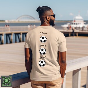 Peut inclure: T-shirt beige avec les noms Oliver, John et Brad imprim&eacute;s au-dessus de graphiques de ballon de football. La personne qui porte le t-shirt est en ext&eacute;rieur, par une journ&eacute;e ensoleill&eacute;e, avec un pont et de l'eau en arri&egrave;re-plan.