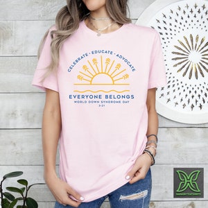May include: Pink t-shirt with a blue and yellow graphic of a sun with rays and the text "Celebrate - Educate - Advocate" above and "Everyone Belongs" below. The text "World Down Syndrome Day 3-21" is also included.