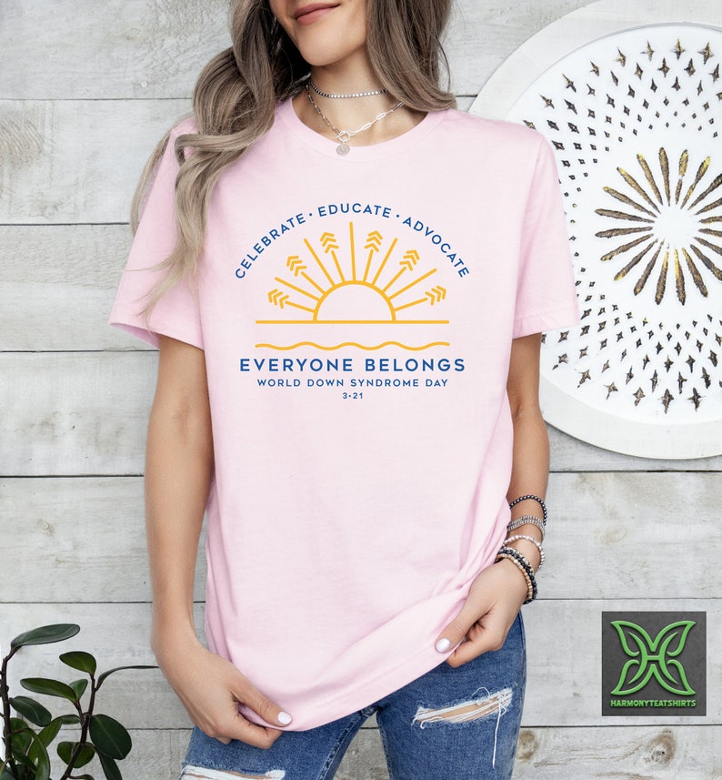 May include: Pink t-shirt with a blue and yellow graphic of a sun with rays and the text "Celebrate - Educate - Advocate" above and "Everyone Belongs" below. The text "World Down Syndrome Day 3-21" is also included.