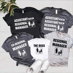 K&ouml;nnte beinhalten: F&uuml;nf T-Shirts in Schwarz, Grau und Wei&szlig; mit wei&szlig;en Aufdrucken im B&uuml;ro-Stil. Auf den Shirts stehen Slogans wie "Assistant Regional Manager" und "The Boss". Die Shirts werden mit wei&szlig;en Turnschuhen pr&auml;sentiert.