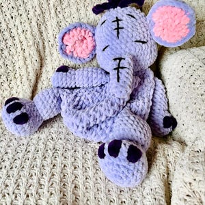 Puede incluir: Un juguete de peluche de elefante lavanda hecho a mano con orejas internas rosas y detalles en morado oscuro en los pies y la cabeza. El elefante está hecho de hilo suave y texturizado y tiene detalles cosidos en negro. Descansa sobre una manta de punto color crema.