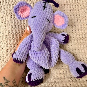 May include: A handmade, plush elephant toy in shades of purple, with pink inner ears and dark purple accents on the feet and trunk. The elephant has closed eyes and a small cross on its forehead.