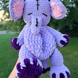 May include: A plush crochet elephant toy made of soft light purple yarn. The elephant features large ears with pink inner sections, dark purple accents on the feet, and a dark purple bow on top. Black yarn stitching details the forehead and trunk.