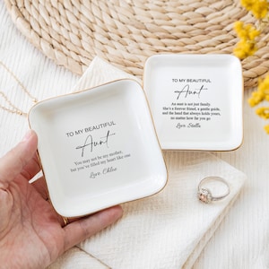 May include: Two white ceramic trinket dishes with gold trim. Each dish has a message for an aunt, with the text "To my beautiful Aunt" and a personalized message below. One dish says "You may not be my mother, but you've filled my heart like one. Love, Chloe." The other dish says "An aunt is not just family. She's a forever friend, a gentle guide, and the hand that always holds yours, no matter how far you go. Love, Stella."