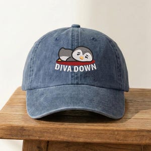 May include: A blue denim baseball cap with a curved brim. The cap features an embroidered design of a cartoon penguin resting on a red cushion, with the words "DIVA DOWN" in white lettering.