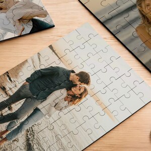 May include: A couple poses on a beach with a sunset in the background. The man is wearing a dark blue shirt and jeans. The woman is wearing a white shirt and jeans. The image is a jigsaw puzzle.