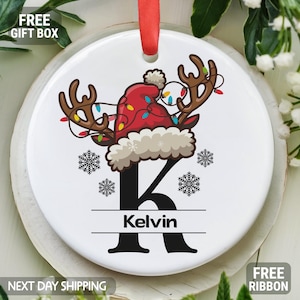 May include: White ceramic ornament with a red and white Santa hat with a reindeer wearing Christmas lights. The ornament has the letter K and the name Kelvin.