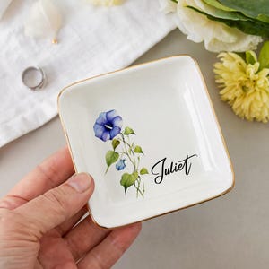 May include: A square, white ceramic trinket dish with gold trim. It features a watercolor design of a blue morning glory flower and the name "Juliet" in black script. The dish is held in a hand.