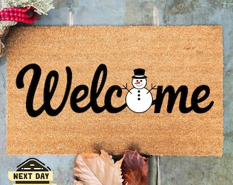 May include: A brown coir doormat with the word "Welcome" in black cursive lettering. A snowman with a black top hat is centered between the "l" and the "c" in the word "Welcome".