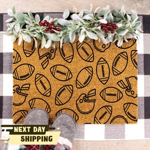 May include: A brown coir doormat with a black and white pattern of footballs and helmets. The doormat is on a black and white chequered surface.