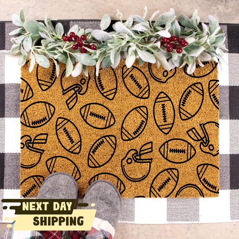 May include: A brown coir doormat with a black and white pattern of footballs and helmets. The doormat is on a black and white chequered surface.