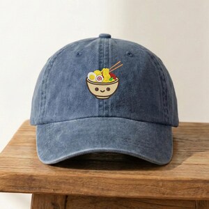 May include: A denim blue baseball cap with a curved brim. The cap features an embroidered design of a smiling bowl of ramen with chopsticks, noodles, and an egg. The cap is sitting on a wooden surface.
