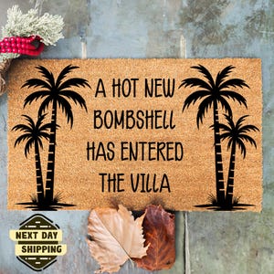 May include: A rectangular tan coir doormat featuring black silhouettes of palm trees on the left and right sides. The text A HOT NEW BOMBSHELL HAS ENTERED THE VILLA is printed in black in the center. The mat has a coarse, natural fiber texture.